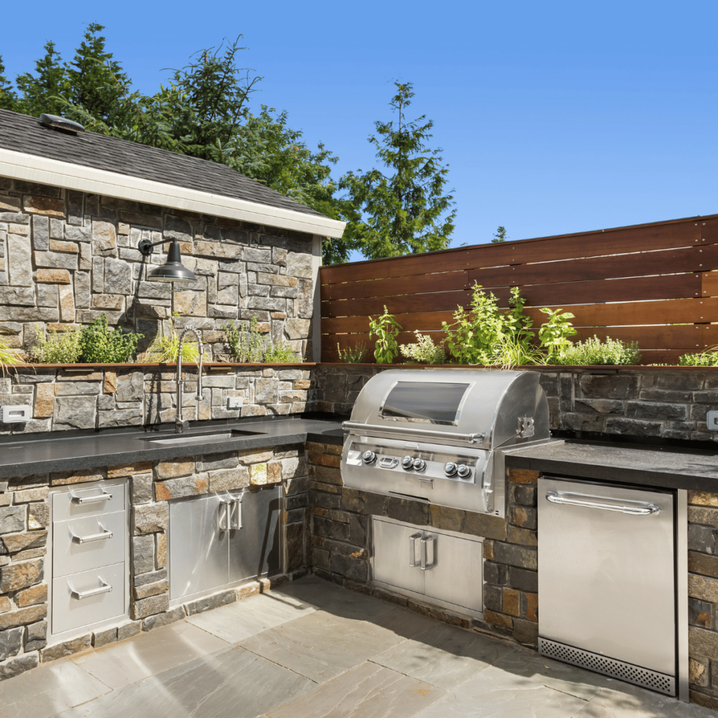 A spacious outdoor kitchen with a built-in grill, sink, and bar seating under a shaded pergola, surrounded by greenery.