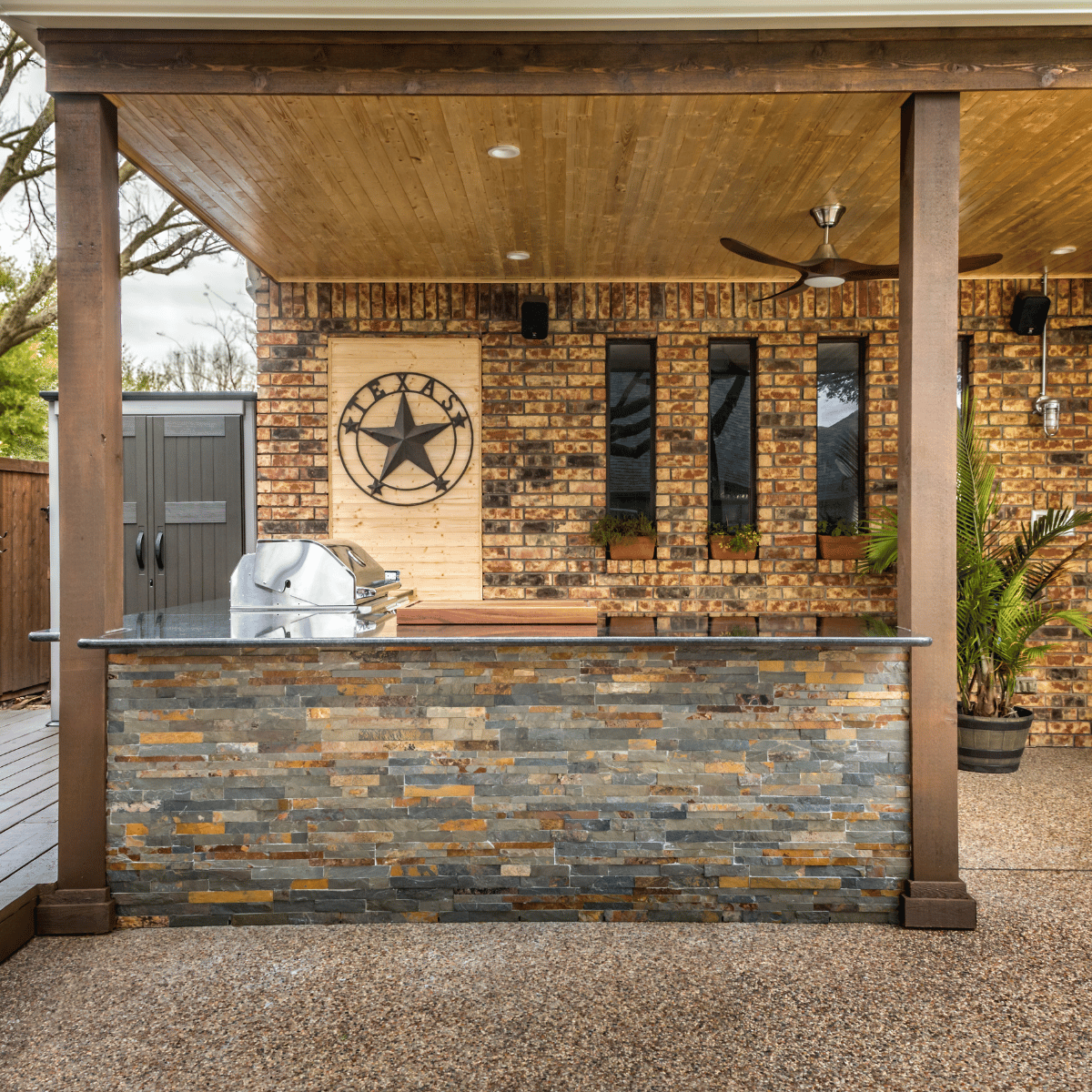 A rustic outdoor kitchen with stone countertops, brick walls, and a built-in stainless steel grill under a wood-paneled ceiling.