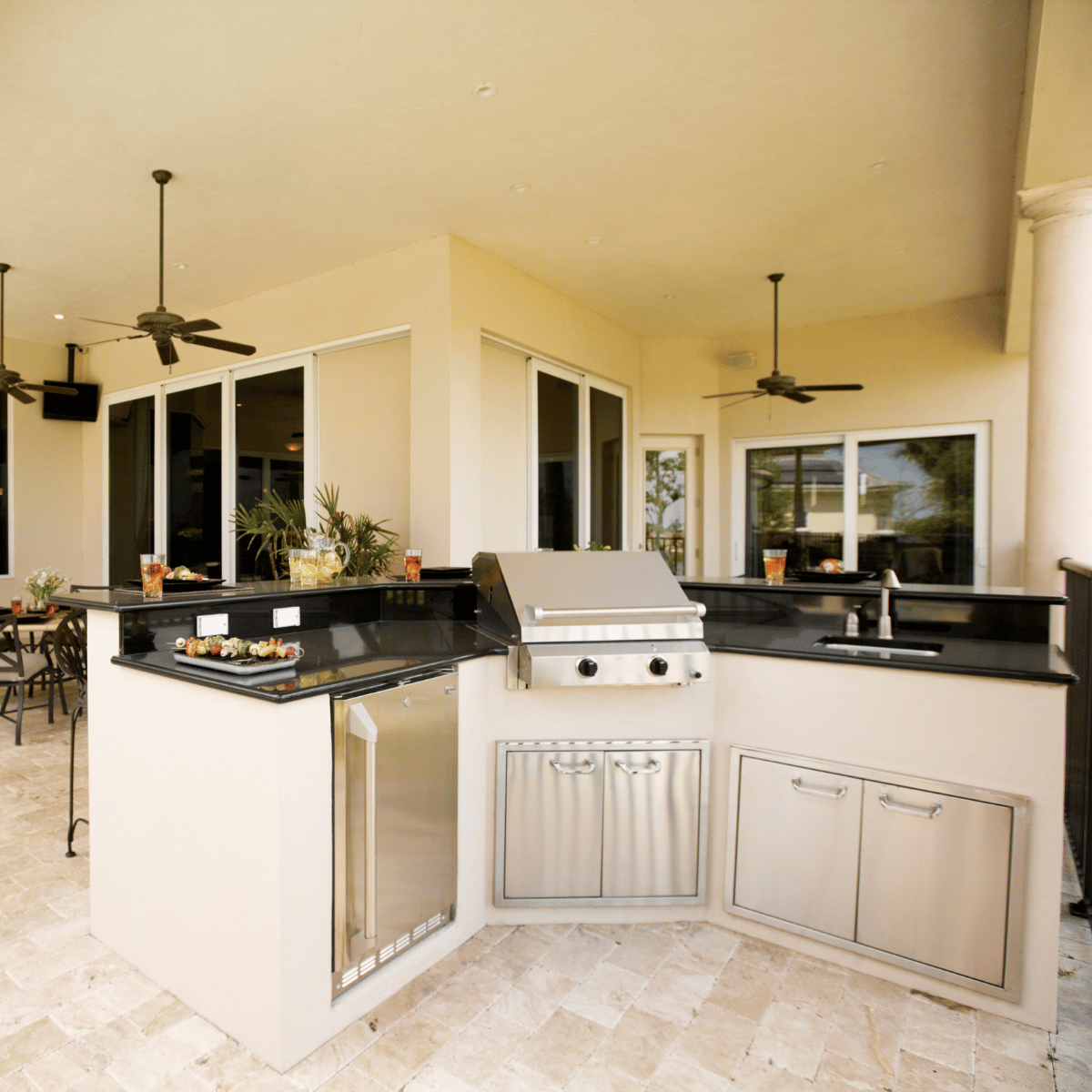 A modern stainless steel outdoor kitchen with a grill, sink, refrigerator, and ample counter space on a tiled patio.